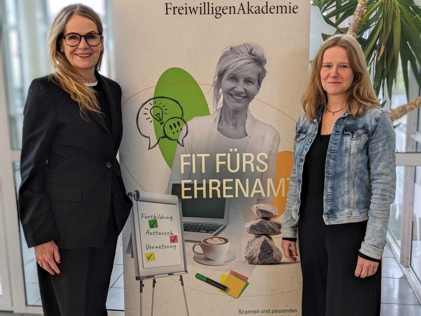 Zwei Frauen stehen neben einem Plakat der FreiwilligenAkademie mit dem Motto „Fit fürs Ehrenamt“ und Checklisten.