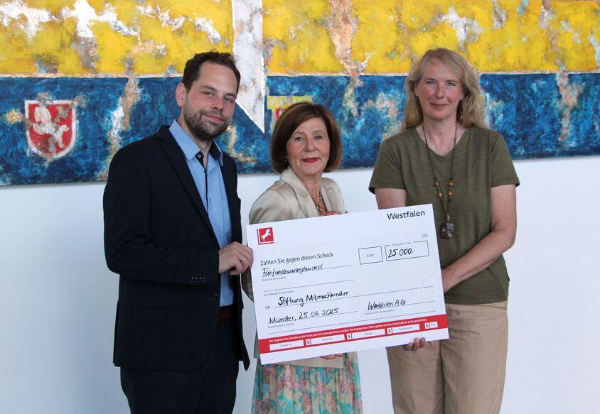 Drei Personen halten einen Scheck über 25.000 Euro vor einem bunten, abstrakten Kunstwerk.